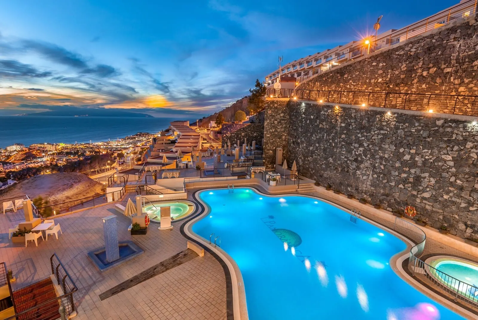 KN Aparthotel Panorámica: A swimming pool with a stone wall and a view of the city at dusk